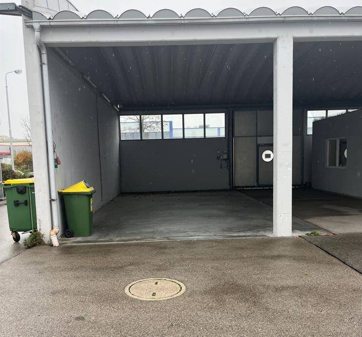 Überdachter Parkbereich oder Ladezone mit Betonboden und Mülltonnen daneben.