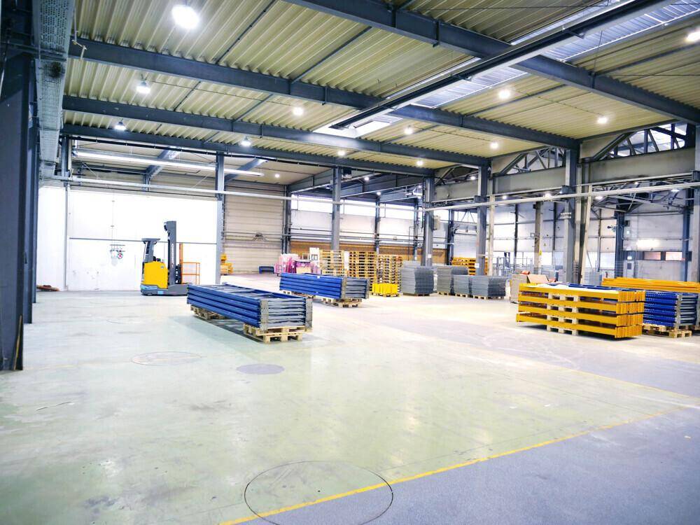 Weitläufige Lagerhalle mit Betonboden, Gabelstapler und gestapelten blauen und gelben Elementen unter Oberlichtern.