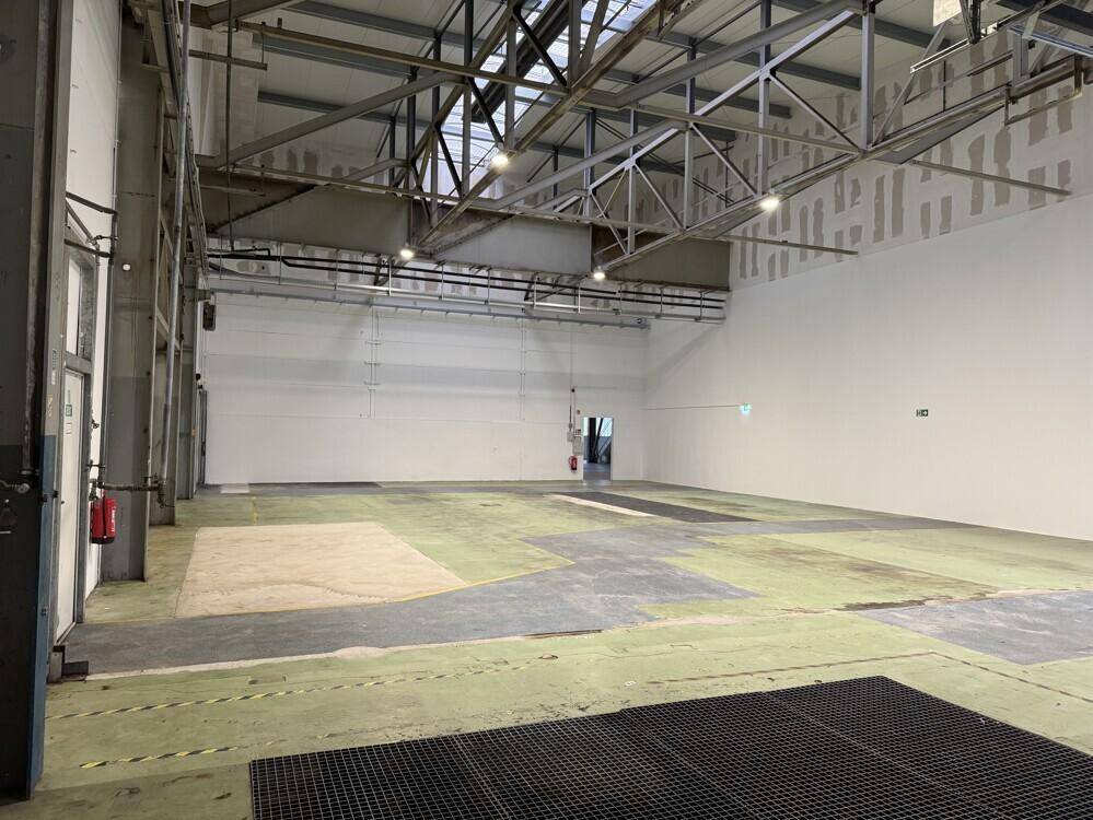 Leere Lagerhalle mit hohen Wänden, sichtbaren Trägern und einem Boden mit verschiedenen Markierungen und Texturen.