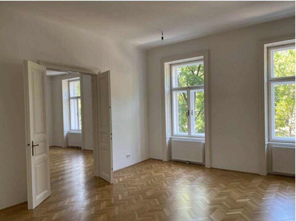 Heller Wohnraum mit Fischgrätparkett, weißen Wänden und zwei großen Fenstern mit Blick ins Grüne.