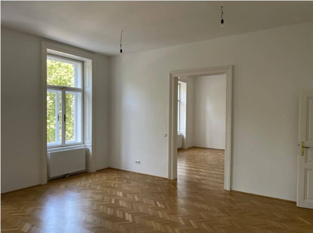 Geräumiger Raum mit Fischgrätparkett und weiß gestrichenen Wänden, mit Zugang zu einem weiteren Zimmer.