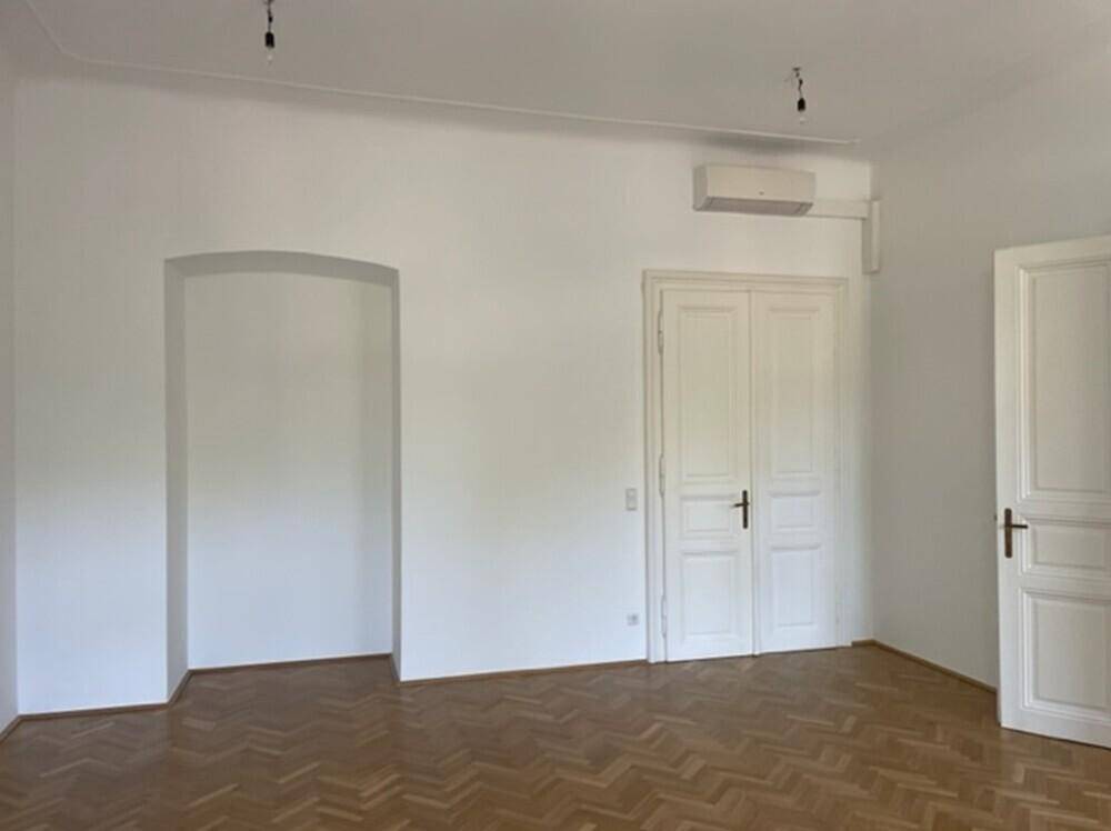 Heller Raum mit Fischgrätparkett, weißen Wänden, einem Bogen und einer Klimaanlage an der Decke.
