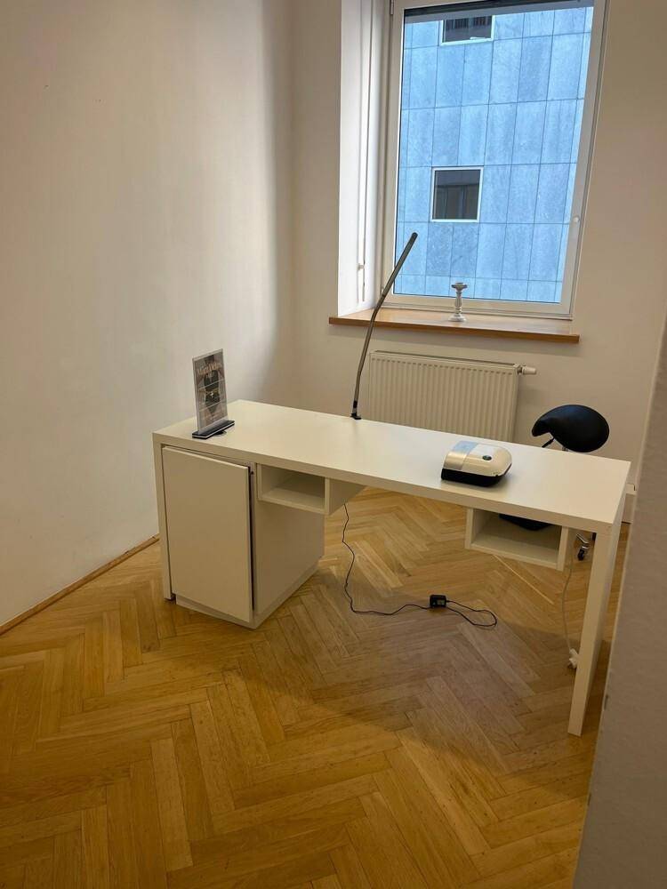 Schlichter Büroraum mit weißem Schreibtisch und Fischgrätparkett, beleuchtet durch ein Fenster mit Ausblick.