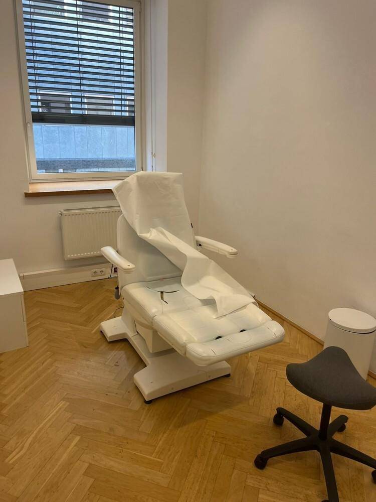 Behandlungsraum mit weißem Stuhl und Fischgrätparkett, Fenster mit Jalousien sorgt für Privatsphäre und Licht.