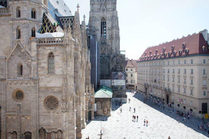 Historische Stadtansicht mit imposantem Dom und belebtem Platz an einem sonnigen Tag, umgeben von Gebäuden.
