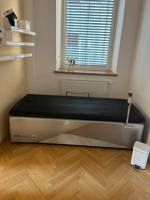 Behandlungsraum mit medizinischem Gerät und Fenster mit Jalousien, ausgestattet mit Holzparkett und weißen Wänden.
