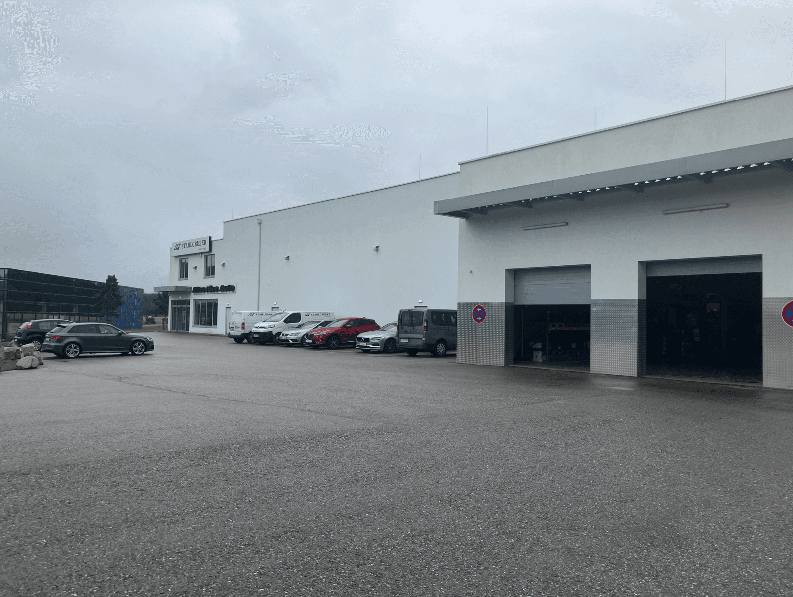 Längsseite des Geschäftsgebäudes mit mehreren geparkten Fahrzeugen und zwei geöffneten Garagentoren für den Zugang.
