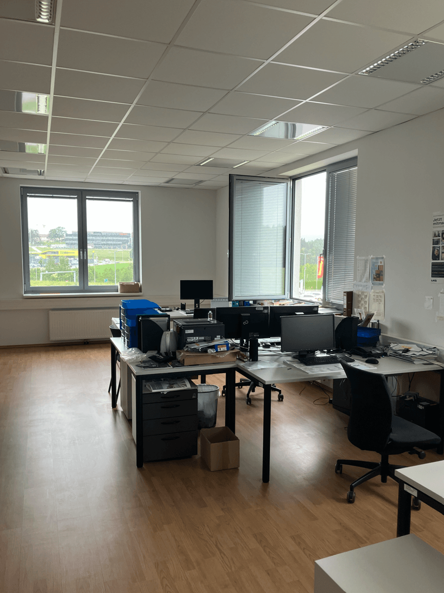 Helles Großraumbüro mit mehreren Arbeitsplätzen, Computern und großen Fenstern, die viel Tageslicht hereinlassen.
