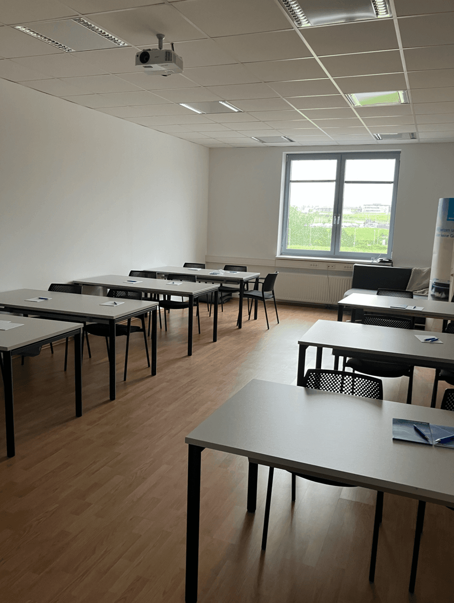 Schulungsraum mit mehreren Tischen und Stühlen, einem Beamer und einem Fenster, ideal für Präsentationen und Meetings.