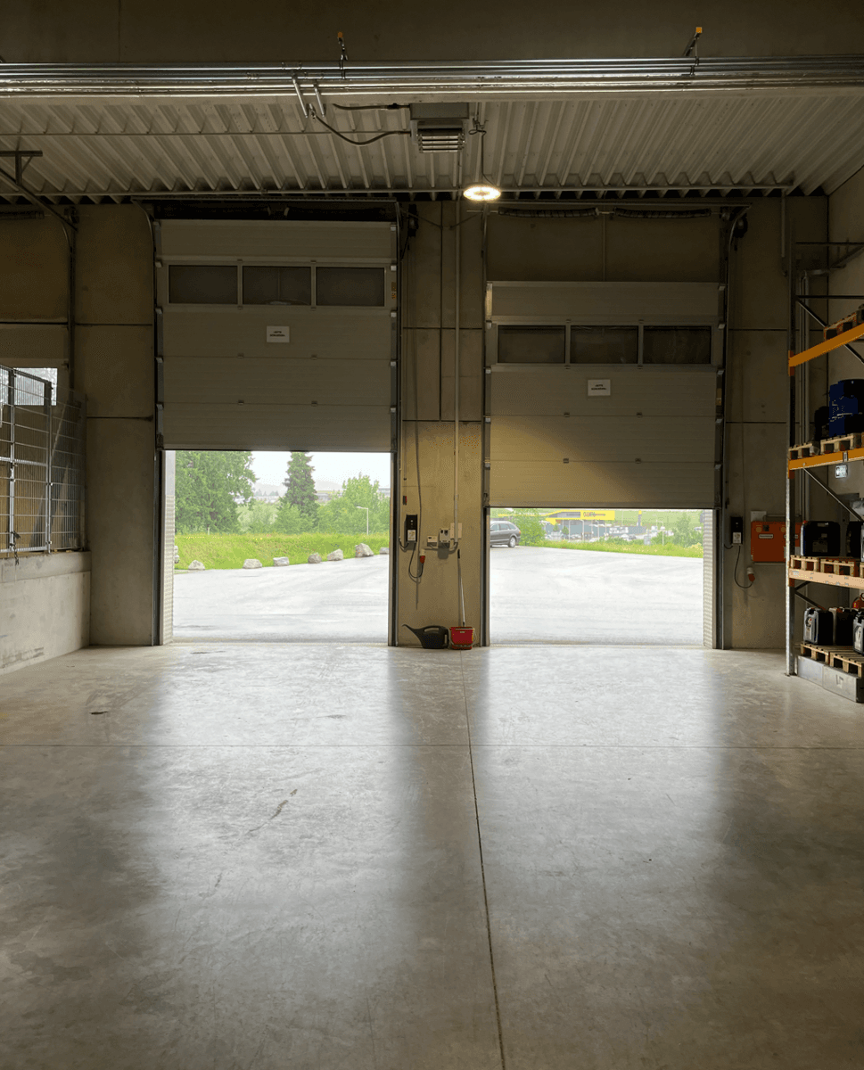 Blick aus einer Lagerhalle durch zwei große, geöffnete Rolltore auf den Außenbereich und die Parkplätze.