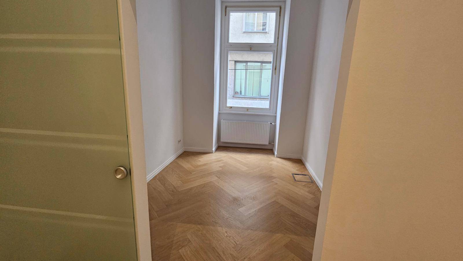 Heller, leerer Raum mit Fischgrätparkettboden, weißen Wänden und einem großen Fenster mit Heizkörper.