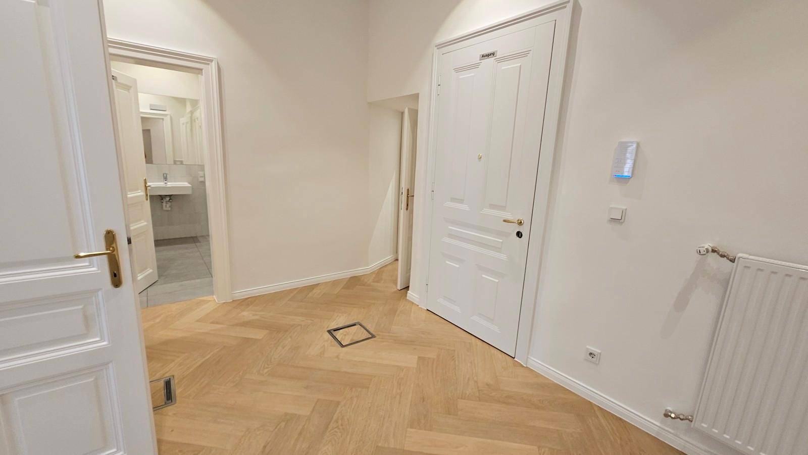 Heller Raum mit Fischgrätparkettboden und weißen Türen, eine davon führt zu einem Badezimmer.