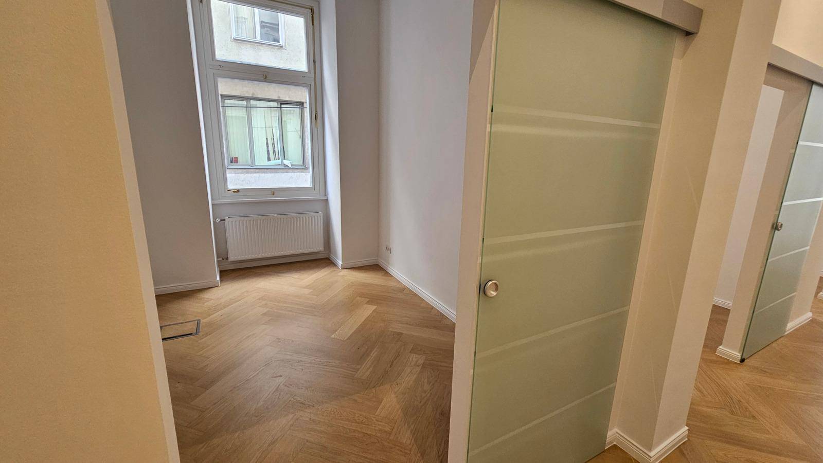 Heller Raum mit Fischgrätparkettboden, großem Fenster und einer modernen Schiebetür aus Milchglas.
