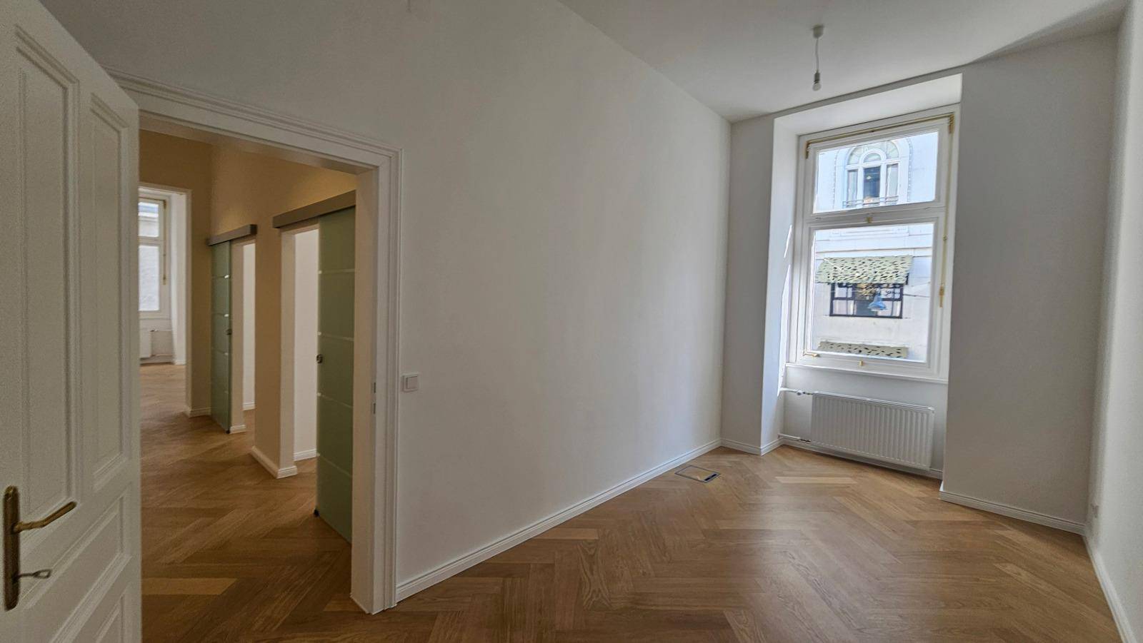 Heller Raum mit Fischgrätparkettboden, großem Fenster und Blick durch offene Türen in angrenzende Zimmer.