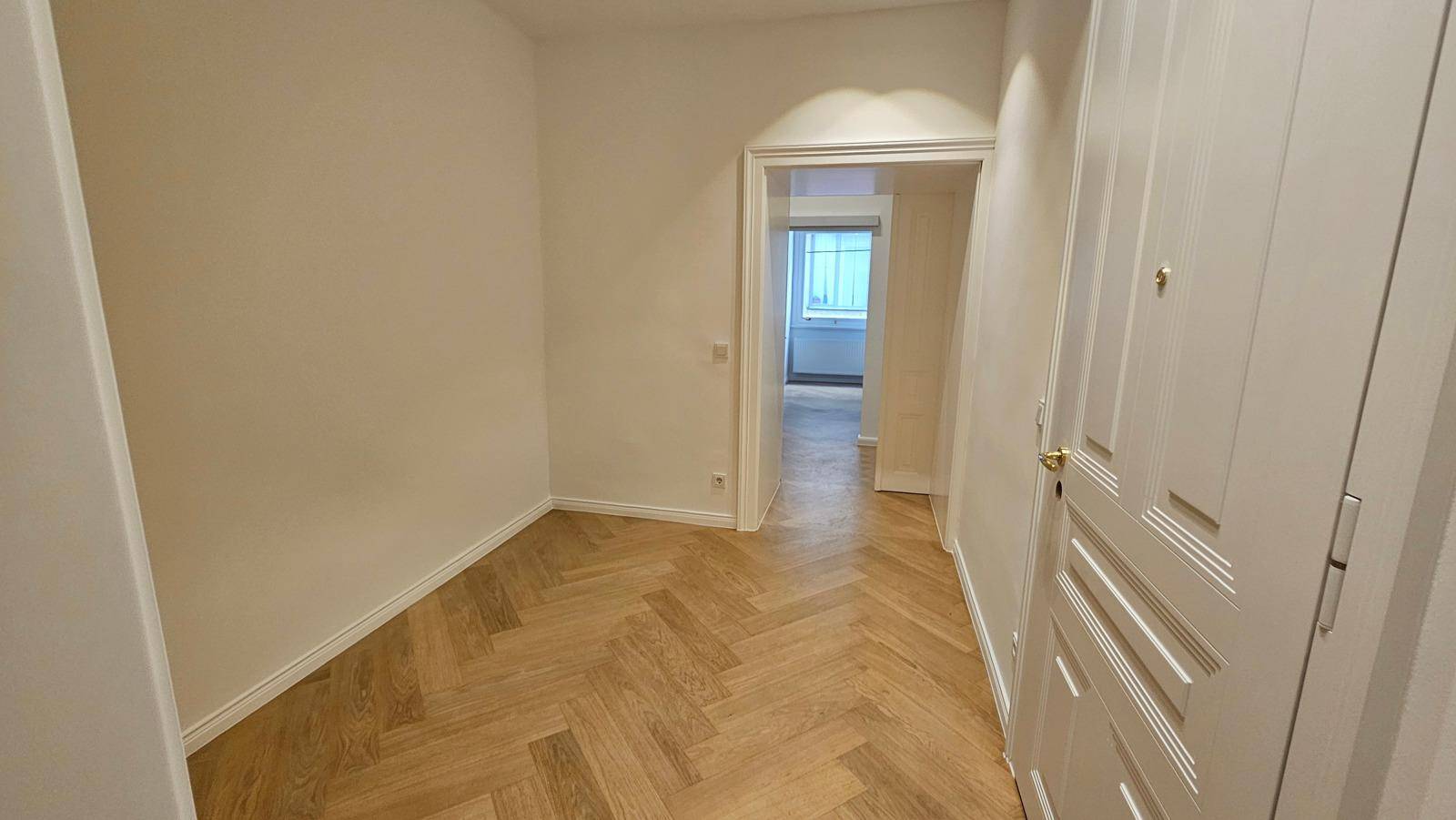 Leerer Raum mit hellem Fischgrätparkettboden und weißen Wänden, der zu einem weiteren Zimmer führt.