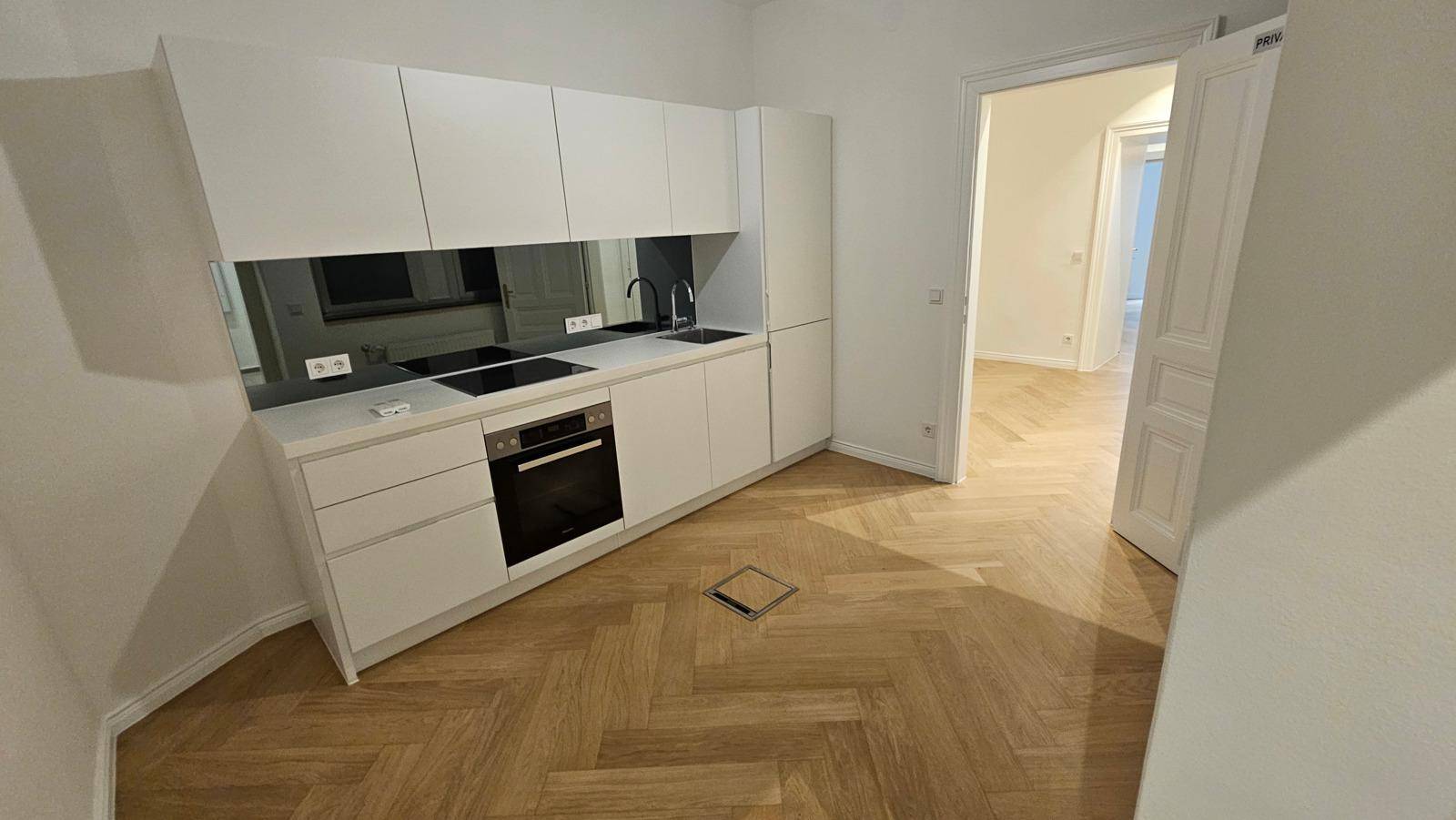 Moderne Küchenzeile mit weißen Schränken, schwarzer Rückwand und integrierten Geräten auf Fischgrätparkett.