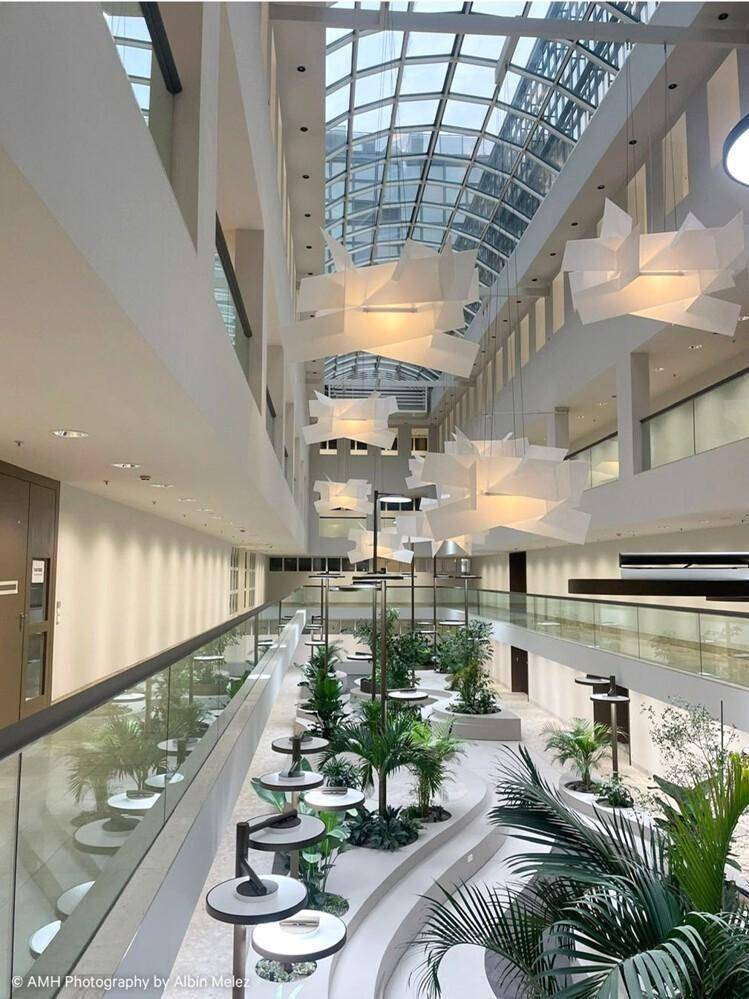 Helles, modernes Atrium mit Glasdach, üppiger Bepflanzung und schwebenden Designleuchten, das eine offene Atmosphäre schafft.