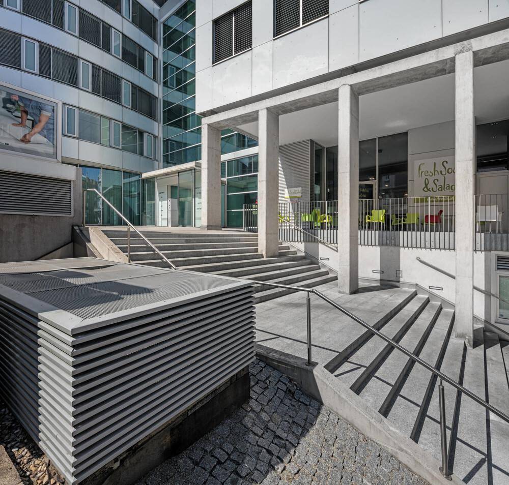 Eingangsbereich eines modernen Bürogebäudes mit Treppen, Rampe und Außensitzgelegenheiten.