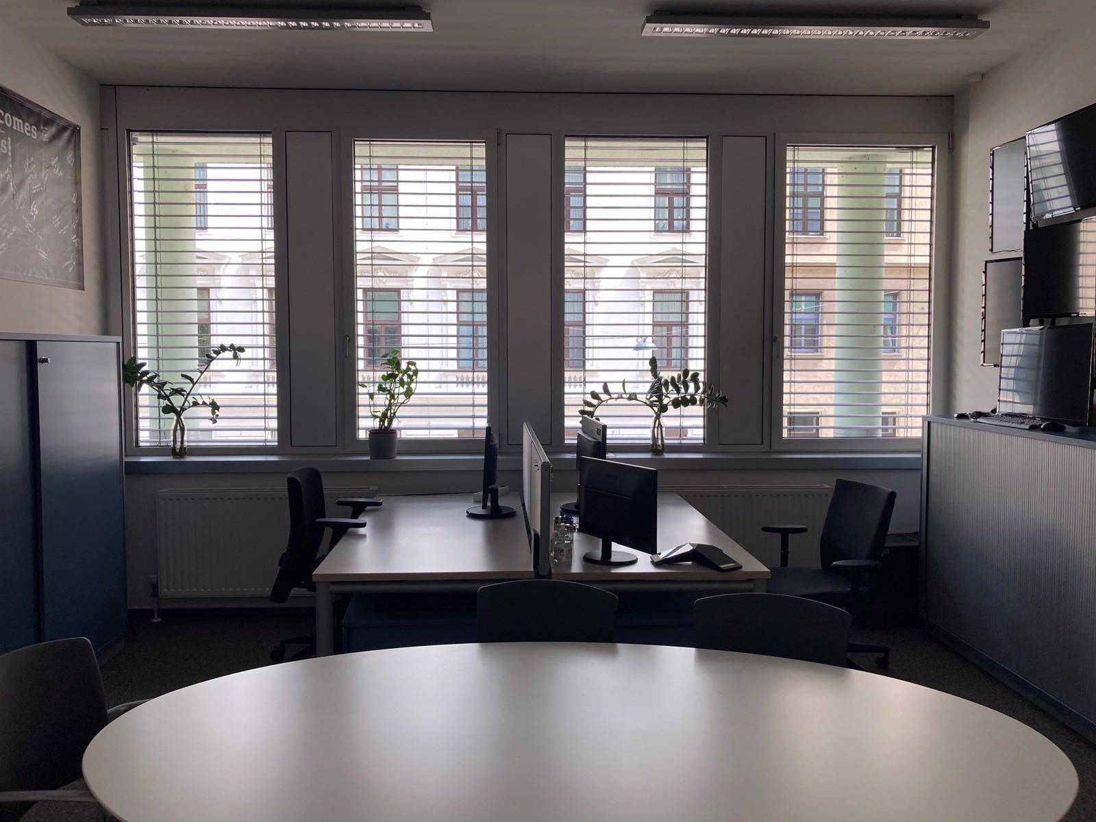 Heller Büroraum mit mehreren Arbeitsplätzen, großen Fenstern und Blick auf die Stadt.