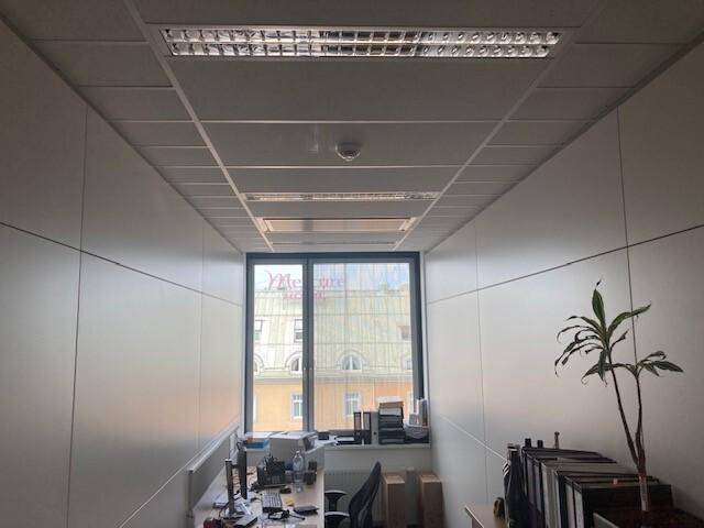 Kleines Büro mit Fenster, Schreibtisch und Deckenbeleuchtung, Blick auf gegenüberliegende Gebäude.