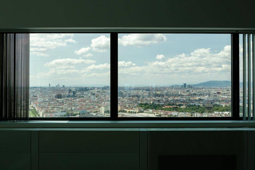 Weitläufiger Stadtblick durch ein großes Fenster mit Jalousien, das eine helle Atmosphäre schafft.