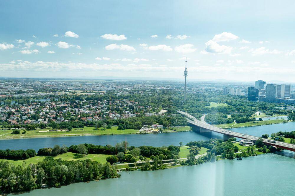 Panoramablick auf die Stadt Wien mit der Donau, Brücken und grünen Ufern unter blauem Himmel.