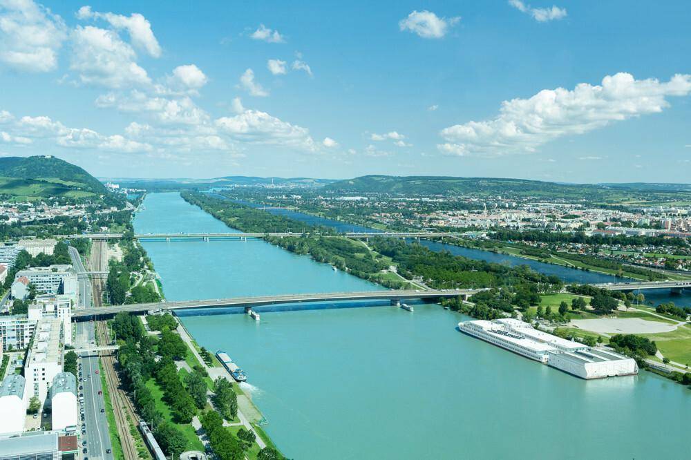 Weitläufiger Blick auf die Donau mit Brücken, Schifffahrt und der umliegenden Stadtlandschaft.