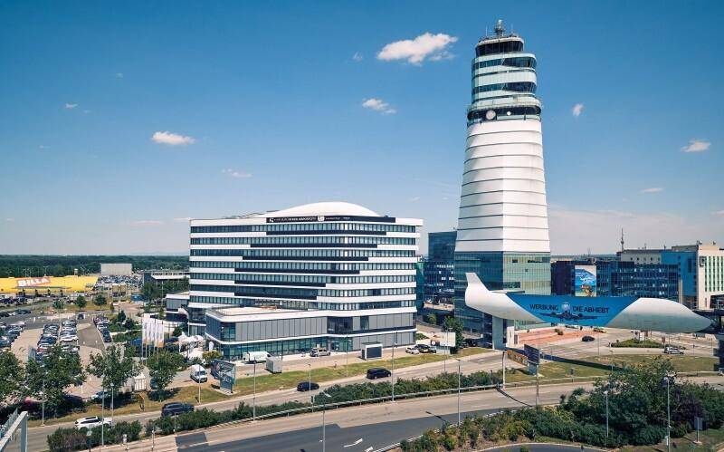 Luftaufnahme des Flughafengeländes mit Kontrollturm, modernem Gebäude und einer Flugzeugskulptur bei Tageslicht.