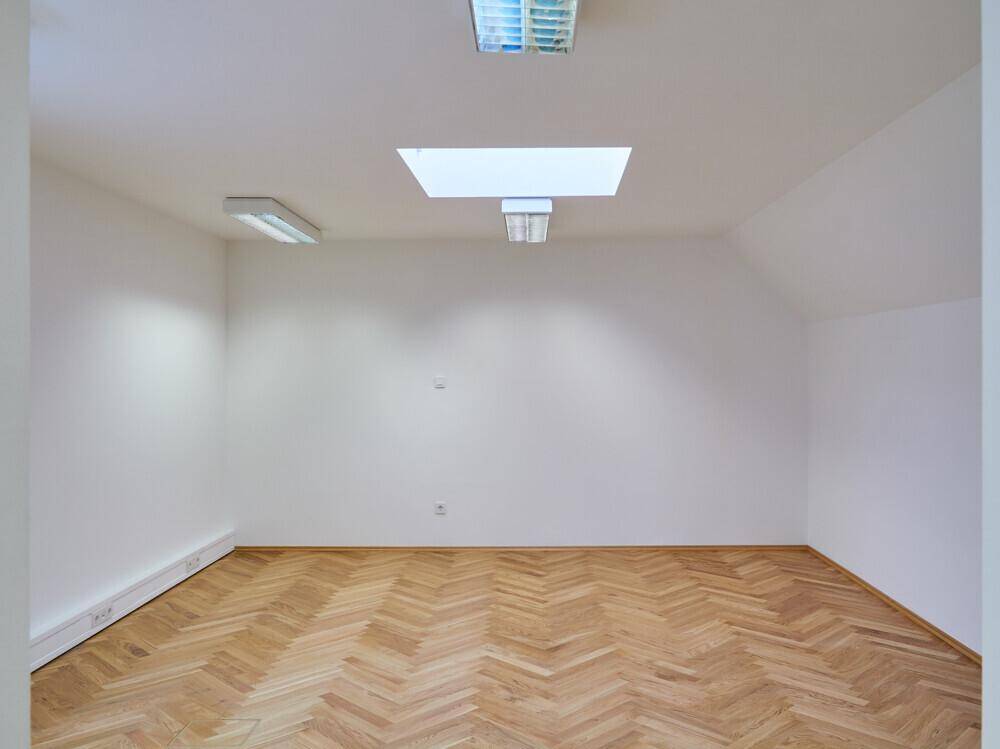 Leerer Büroraum mit Fischgrätparkett, weiß gestrichenen Wänden und einem rechteckigen Dachfenster.