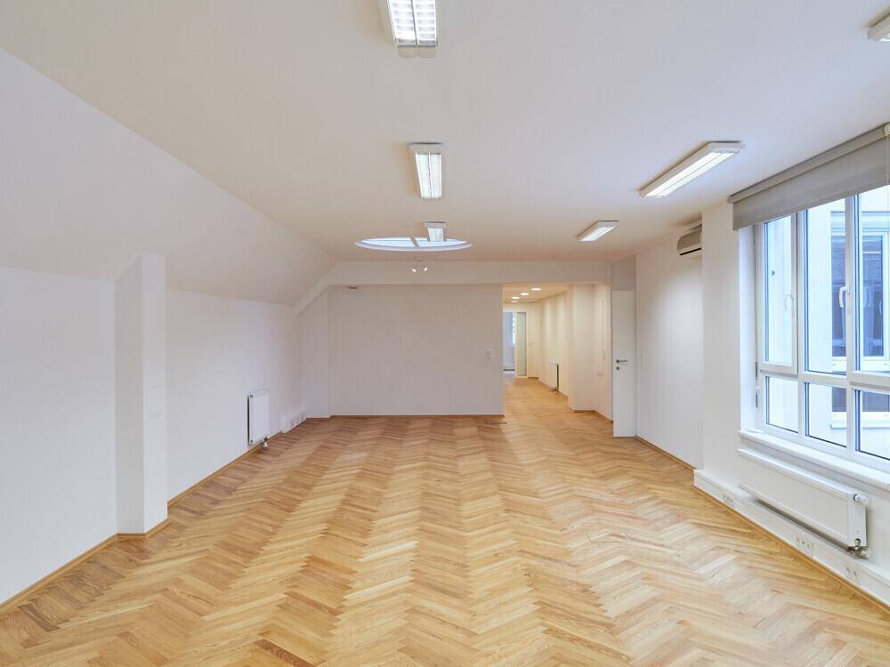 Langer, heller Büroraum mit Fischgrätparkett, Dachfenster und Zugang zu weiteren Räumen.