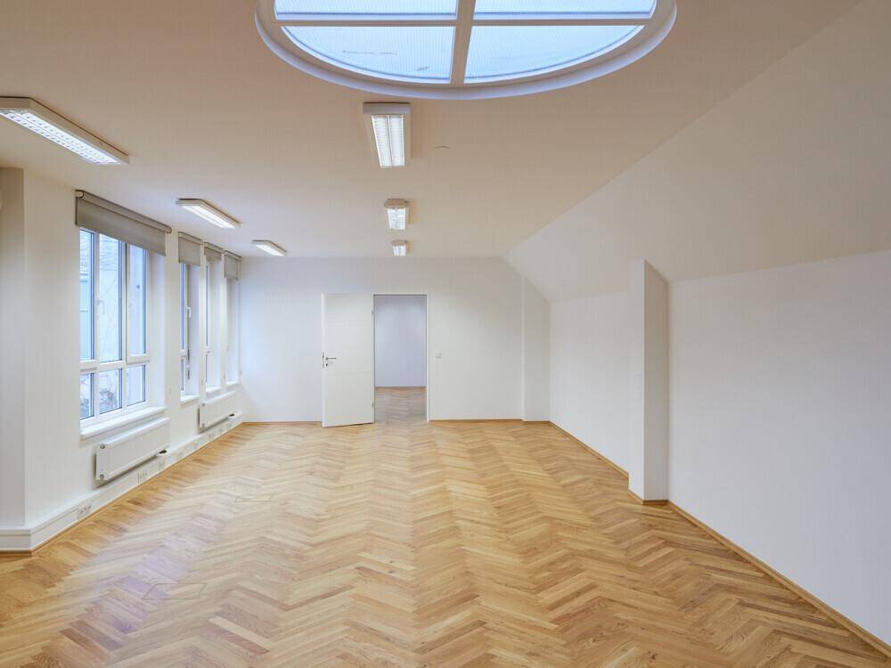 Heller Büroraum mit Fischgrätparkett, mehreren Fenstern und einem markanten ovalen Dachfenster.