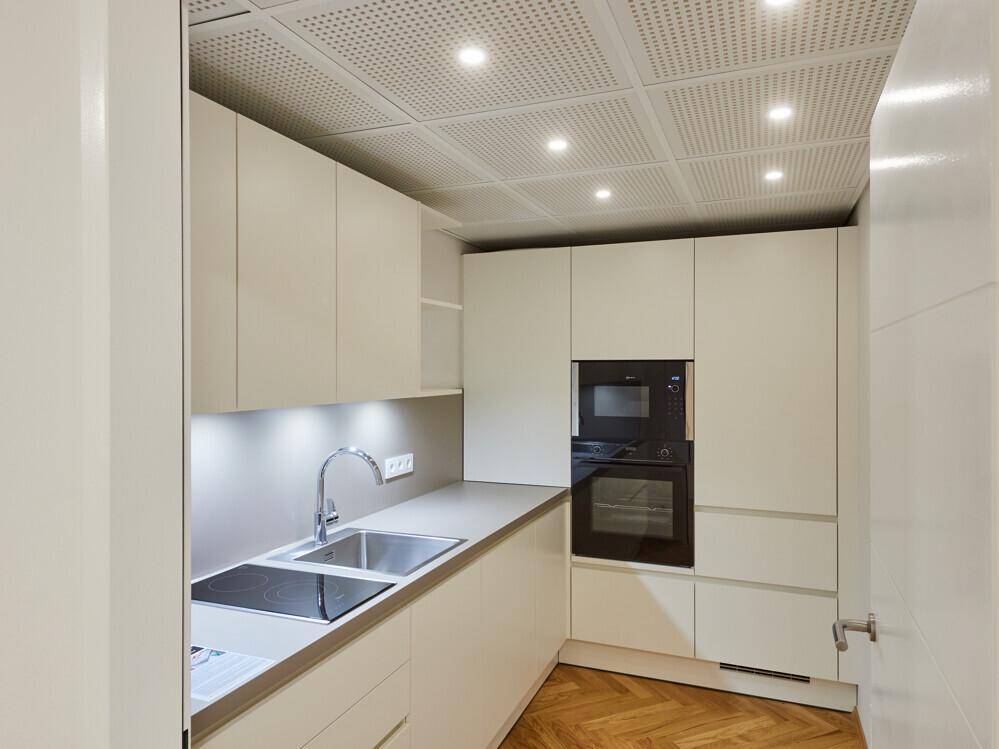 Moderne Einbauküche mit hellen Schränken, integrierten Geräten und perforierter Deckenverkleidung.