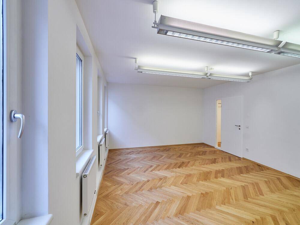 Heller Büroraum mit Fischgrätparkett, großen Fenstern und Blick auf die umliegenden Gebäude.