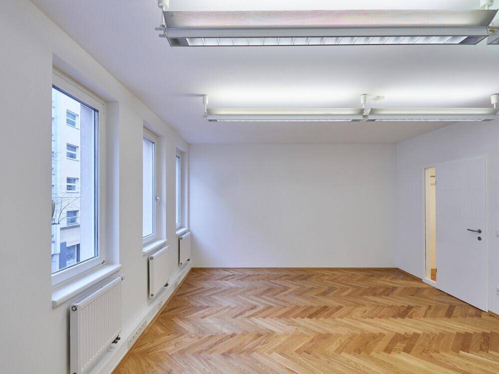 Geräumiger, heller Büroraum mit Fischgrätparkett und großen Fenstern, die viel Licht spenden.