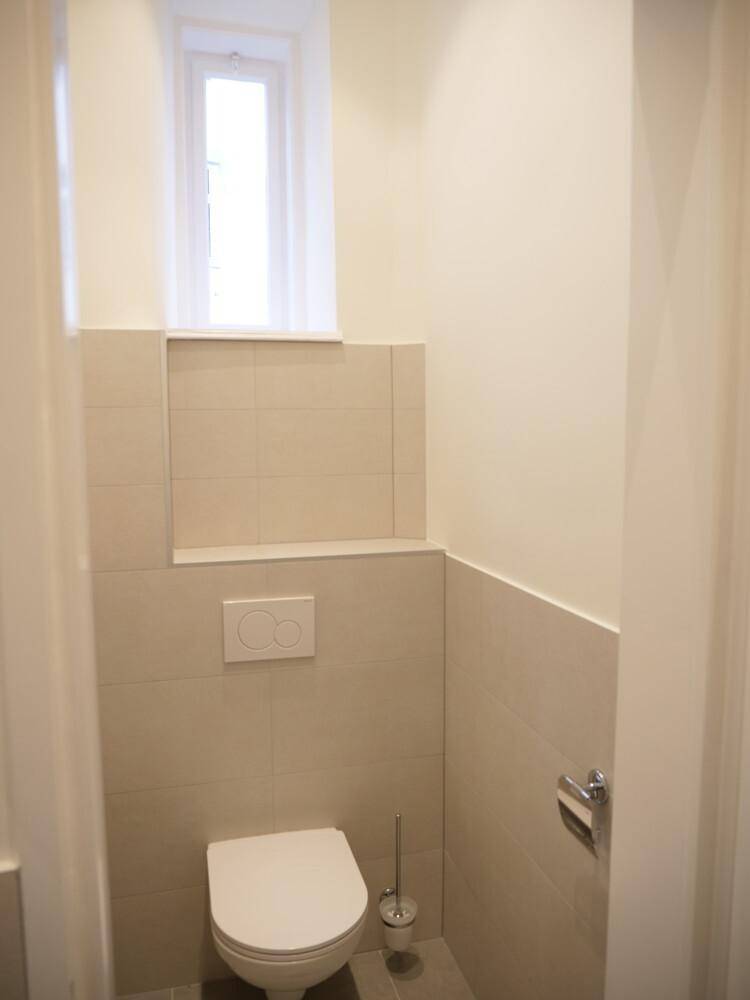 Helles Gäste-WC mit wandhängender Toilette, beigefarbenen Fliesen und einem kleinen Fenster.