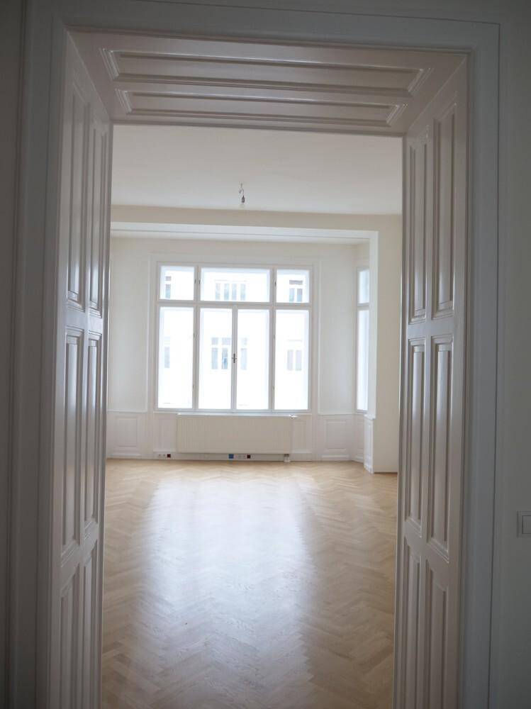 Blick durch eine verzierte Doppeltür in einen hellen Raum mit Fischgrätparkett und Erkerfenster.