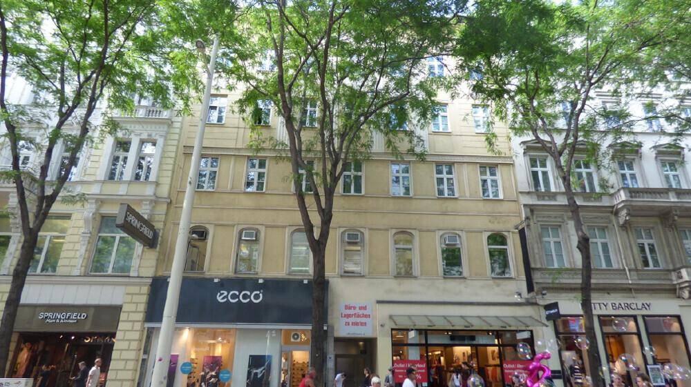 Historische Gebäudefassade mit Geschäften im Erdgeschoss und Bäumen entlang der Straße.
