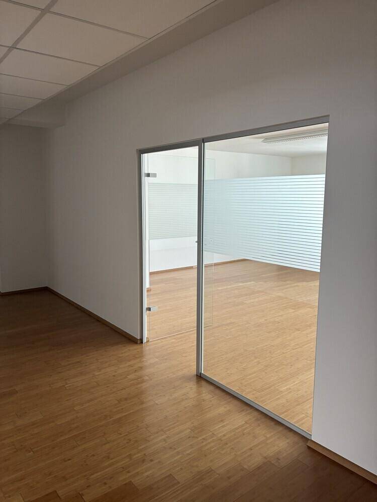 Heller Büroraum mit Holzboden und modernen Glastüren, die zu einem weiteren Bereich führen.