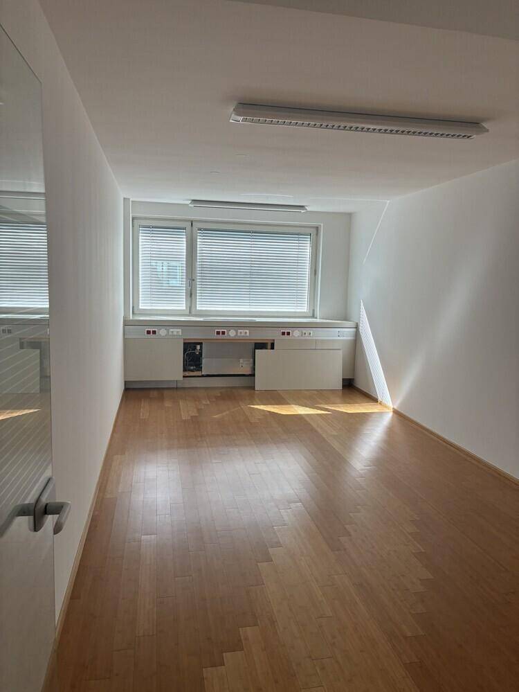 Büroraum mit Holzboden, Fenster mit Jalousien und integrierten Schränken unter der Fensterbank.