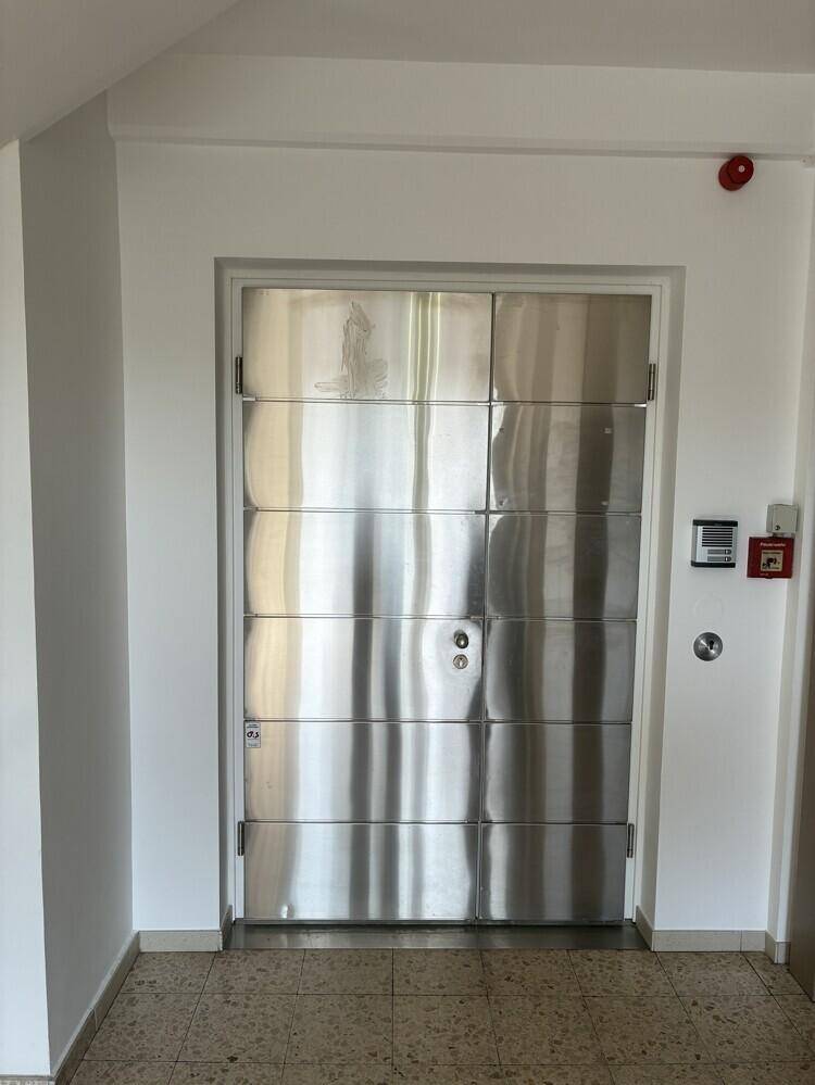 Robuste Metalltür in einem Flur mit Fliesenboden und Sicherheitseinrichtungen an der Wand.