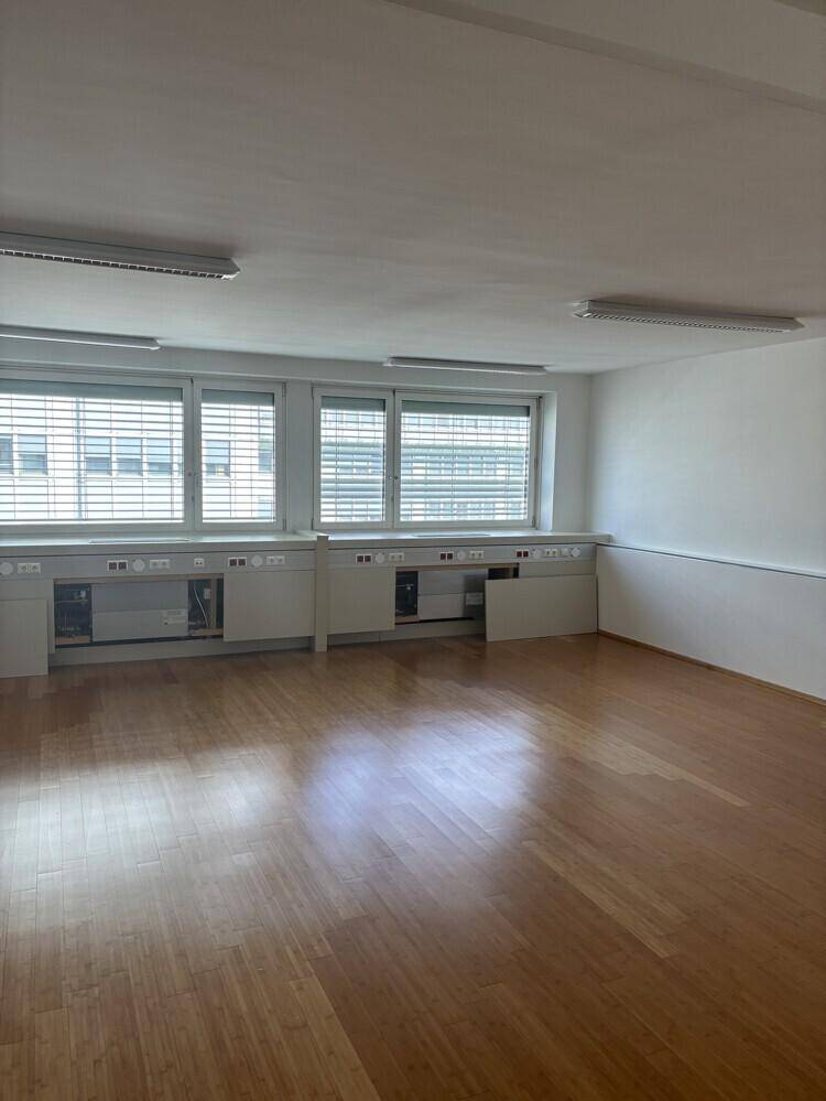 Großer Büroraum mit Holzboden, drei Fenstern mit Jalousien und integrierten Unterschränken.