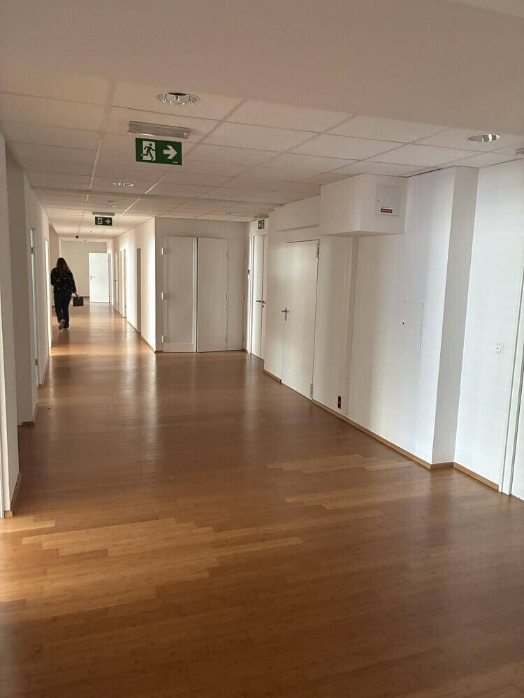Langer Bürogang mit Holzboden, weißen Wänden und Deckenplatten, beleuchtet von Leuchtstoffröhren.