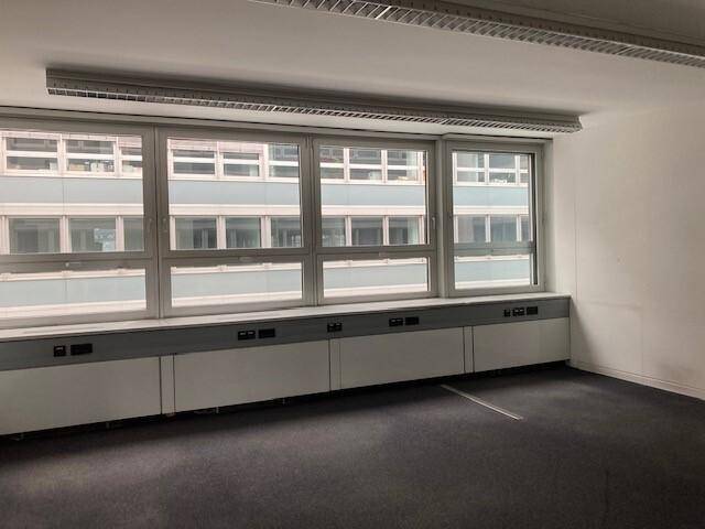 Leerer Büroraum mit dunklem Teppichboden und großen Fenstern, die viel Licht hereinlassen.