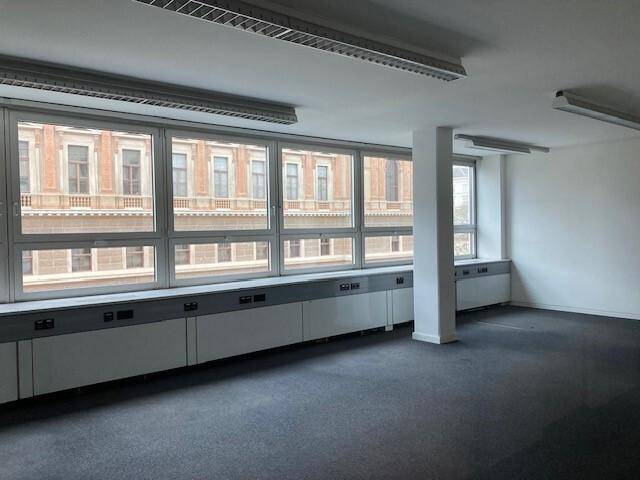 Leerer Büroraum mit dunklem Teppichboden und großen Fenstern mit Blick auf eine Gebäudefassade.