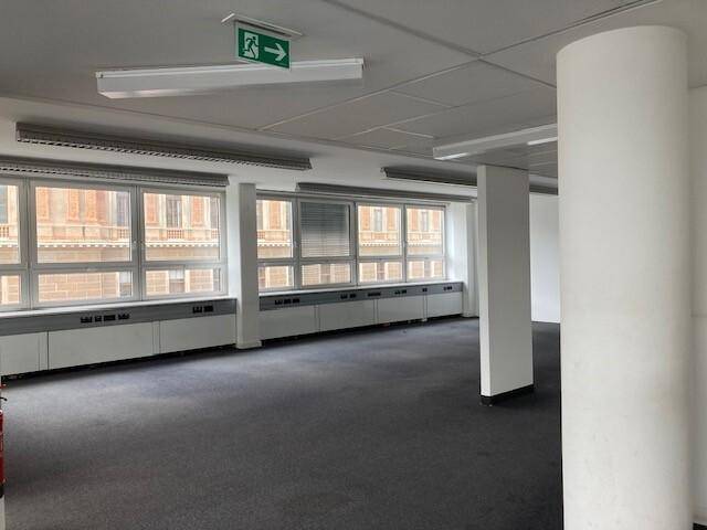 Großer, leerer Bürobereich mit dunklem Teppichboden, Säulen und vielen Fenstern.