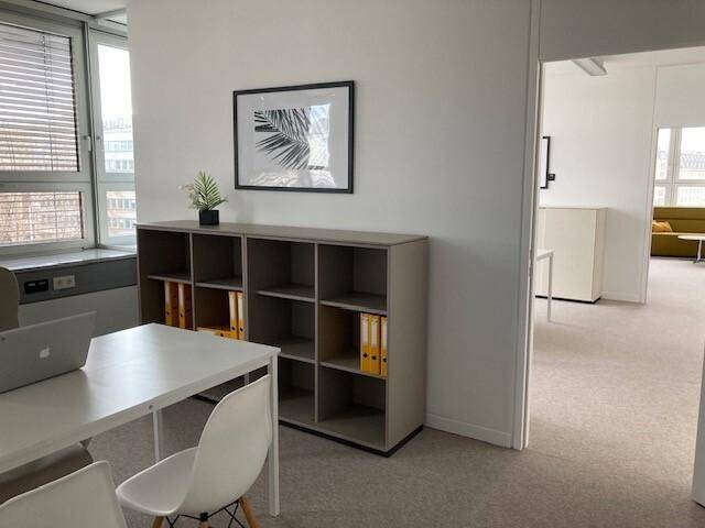 Helles Büro mit Schreibtisch, Stuhl und einem Regal, ausgestattet mit Teppichboden.