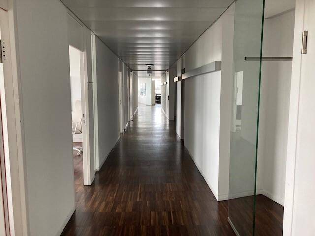 Langer Büroflur mit dunklem Holzfußboden und weißen Wänden, der zu weiteren Räumen führt.