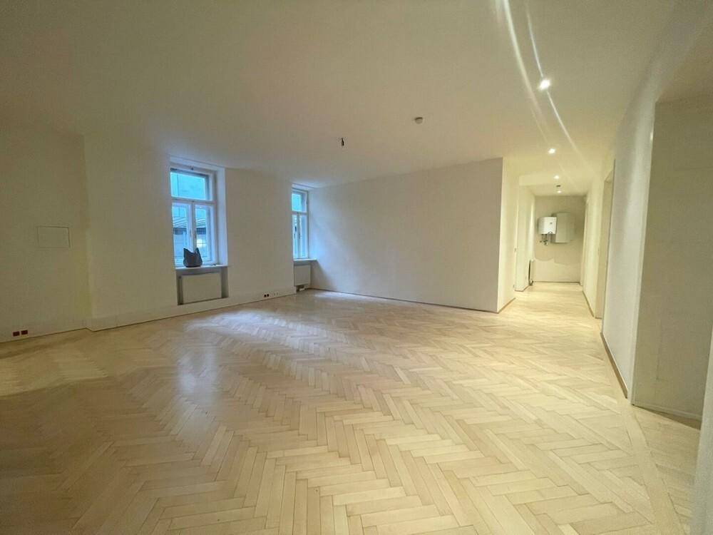 Geräumiger, leerer Wohnbereich mit hellem Fischgrätparkett und zwei Fenstern, die den Raum erhellen.