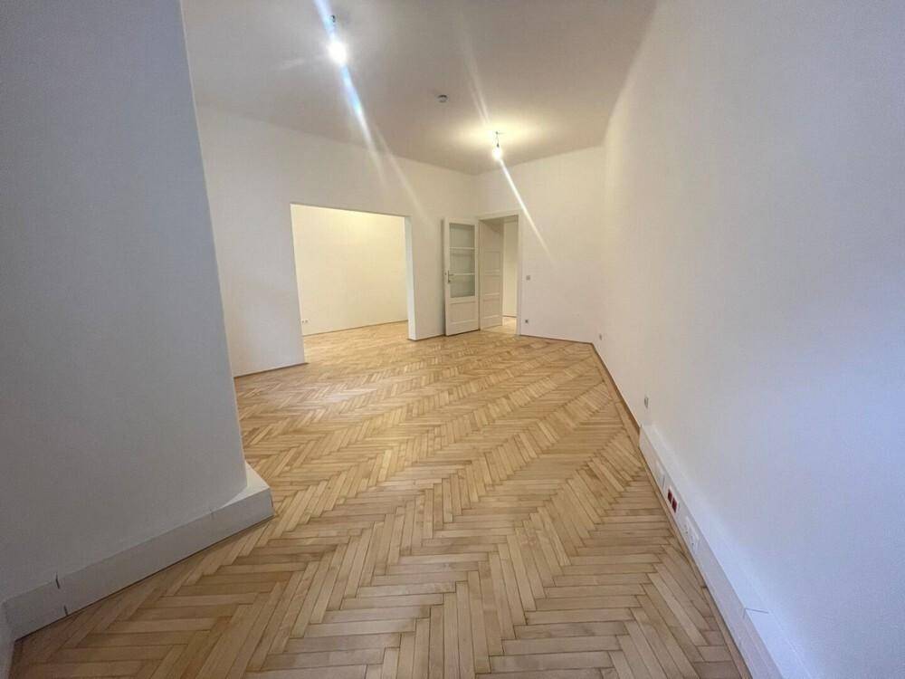 Heller Wohnraum mit Fischgrätparkett und einer offenen Tür, die zu einem weiteren Zimmer führt.
