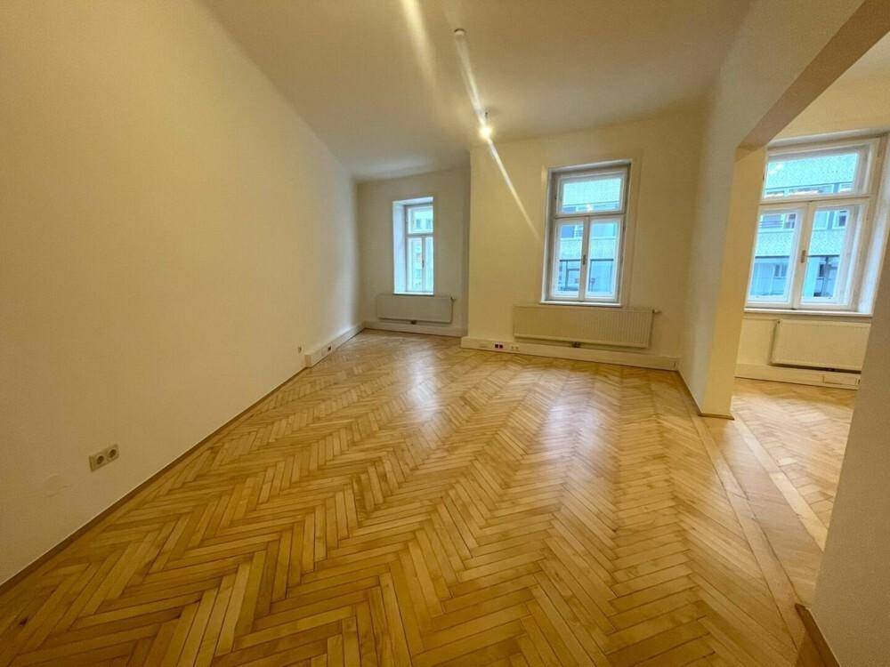 Geräumiger, heller Wohnbereich mit glänzendem Fischgrätparkett und mehreren Fenstern für viel Tageslicht.